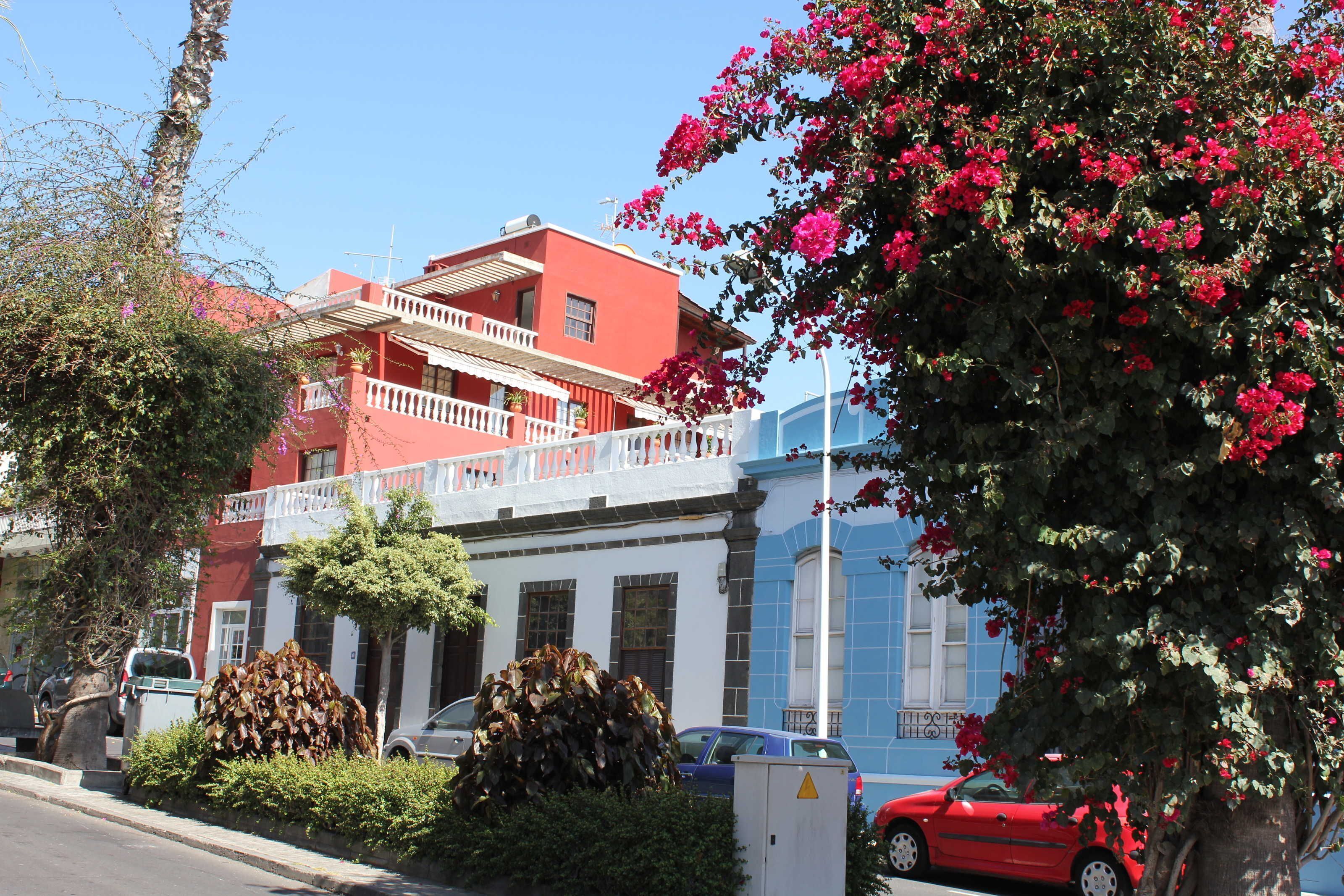A red building with a balcony, surrounded by greenery and parked cars.