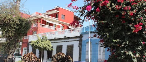 A red building with a balcony, surrounded by greenery and parked cars.