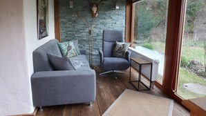 Une pièce chaleureuse avec un canapé et un fauteuil gris, un mur en pierre et une vue sur l’extérieur.
