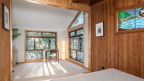 A room with a bed, a desk, and large windows offering a view of trees.