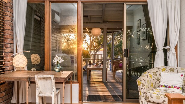 A wooden deck with a table and chairs, a sofa, and a glass door leading to an outdoor area with a bench and a tree.