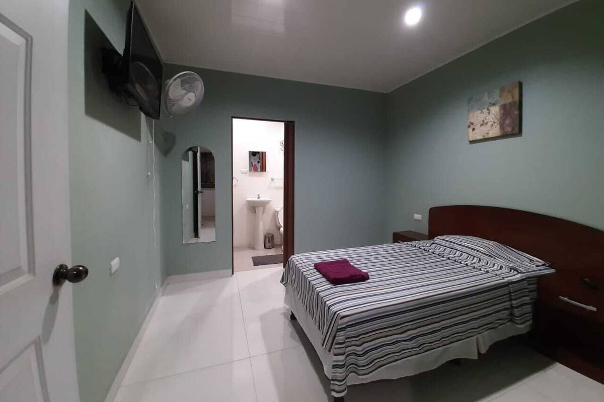 A bedroom with a bed, a bedside table, a wall-mounted TV, and a door leading to a bathroom.