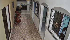Um corredor com chão em mosaico, cadeiras de madeira, área de cozinha com geladeira e janela com grade decorativa.