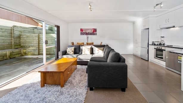 A modern kitchen with stainless steel appliances, a dining area with a sofa and coffee table, and a sliding glass door leading to a balcony.