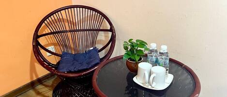 A room with a round glass table, two wicker chairs, a potted plant, and two bottles of water.