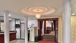 Un vestíbulo de hotel con una recepción, un reloj, una planta, una escalera y un tapete de bienvenida rojo.