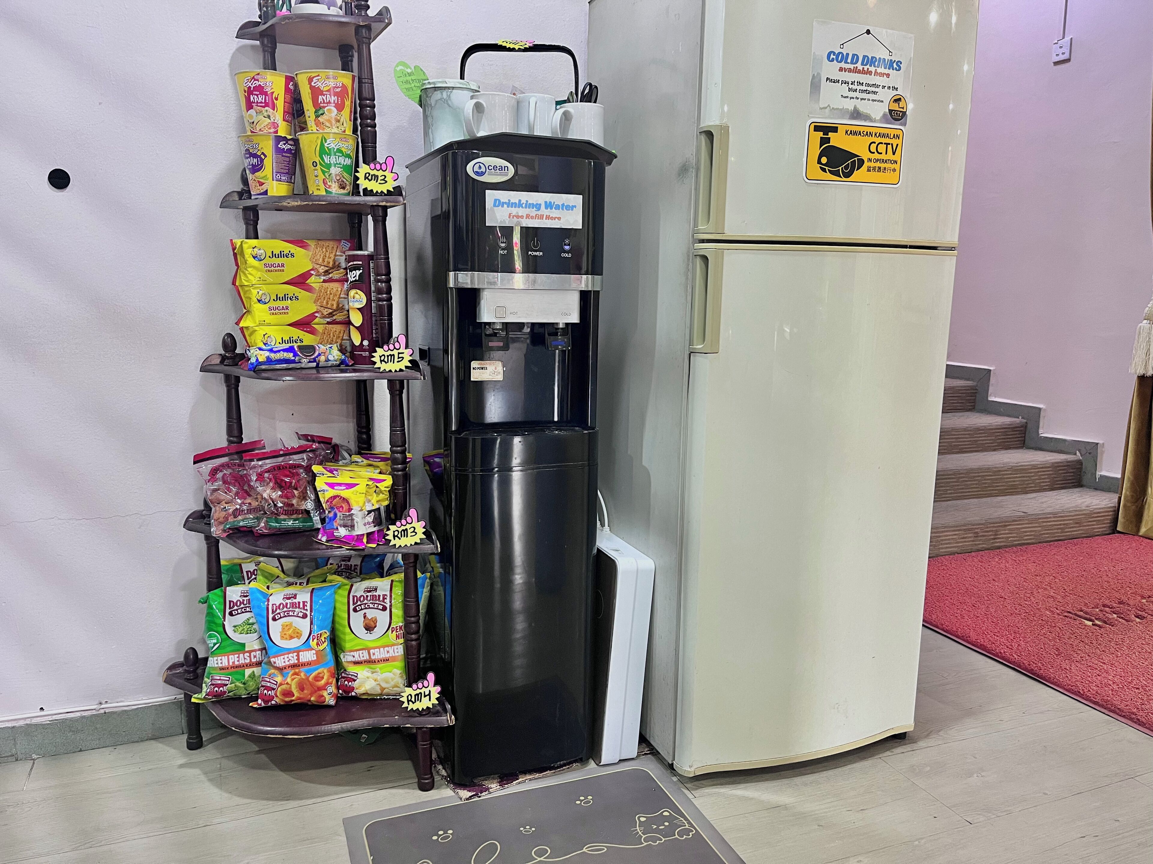 Dispensador de água por autoatendimento, estantes com lanches e uma geladeira em um quarto.