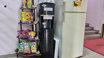 Dispensador de água por autoatendimento, estantes com lanches e uma geladeira em um quarto.
