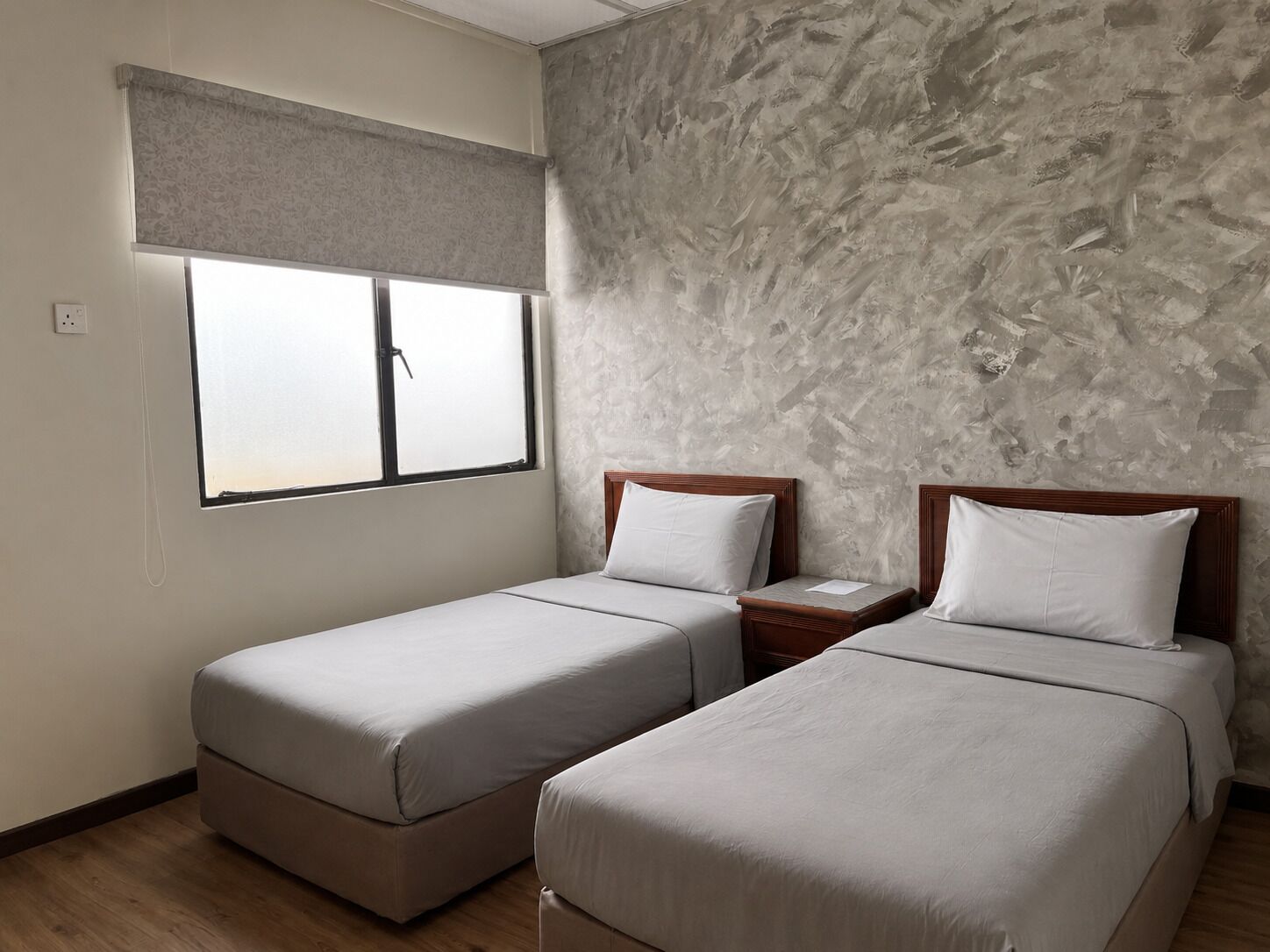 A room with two single beds, a window with a blind, a textured wall, and a wooden floor.