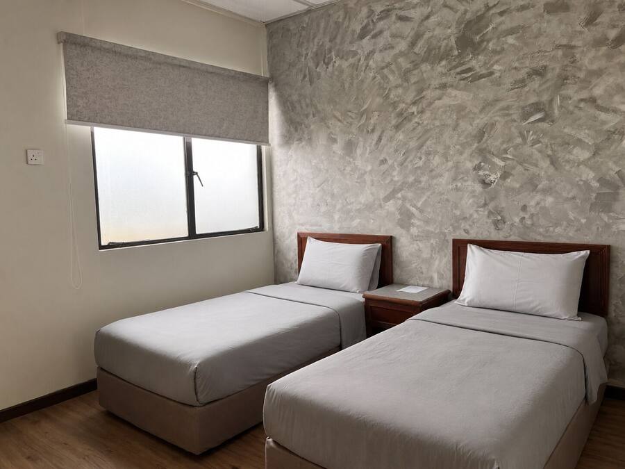 A room with two single beds, a window with a blind, a textured wall, and a wooden floor.