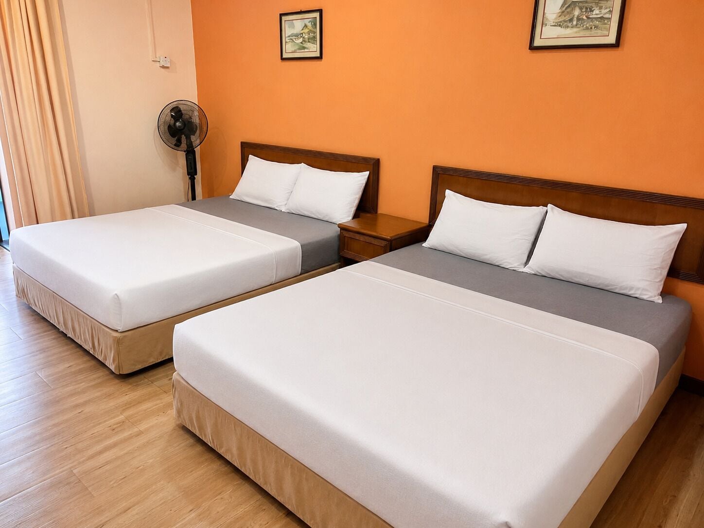 A hotel room with two beds, wooden headboards, a bedside table, a wall-mounted fan, and two framed pictures.