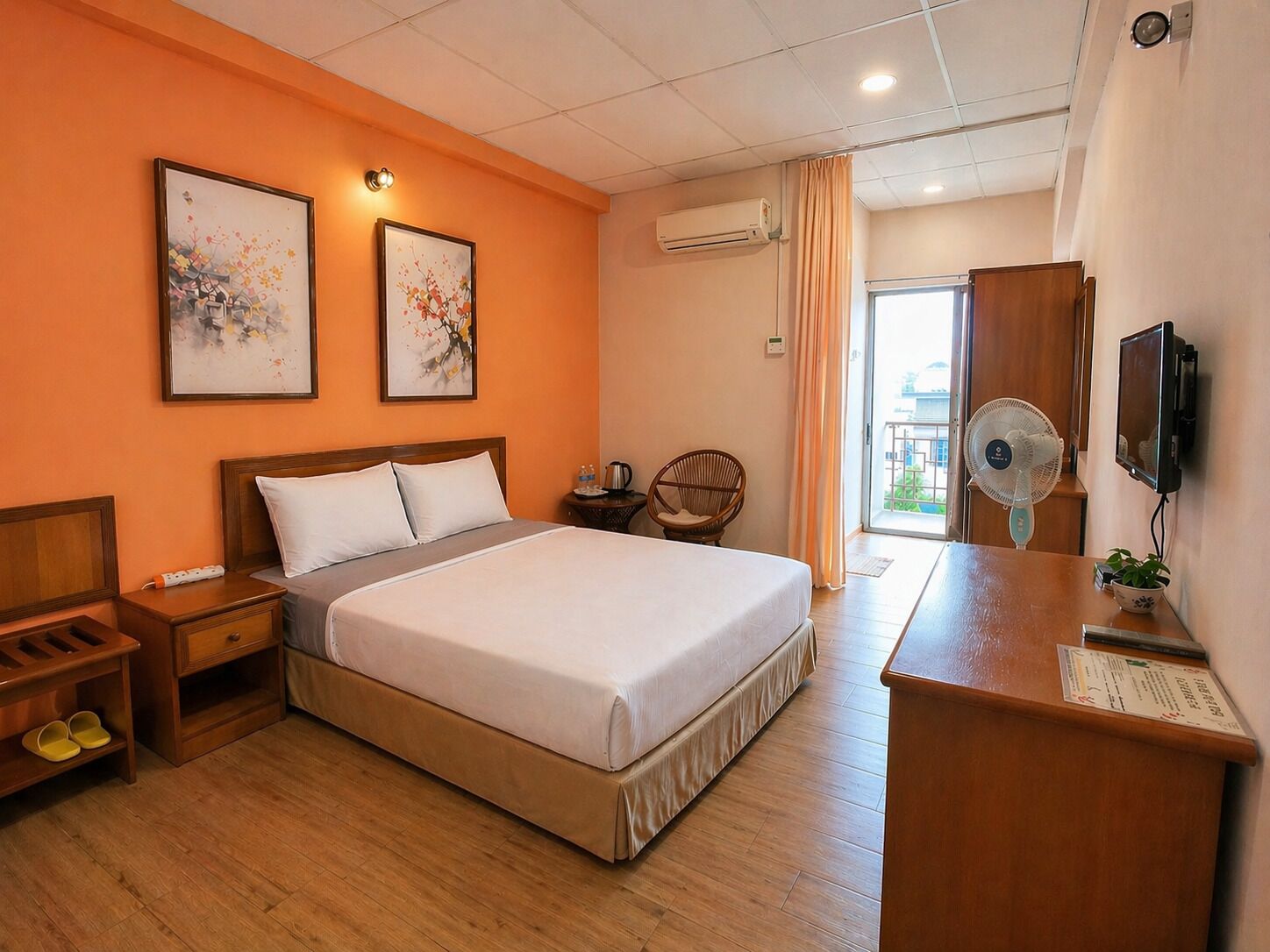 Habitación de hotel con cama, mesitas de noche de madera, un escritorio con silla, televisor, aire acondicionado y dos cuadros en la pared.