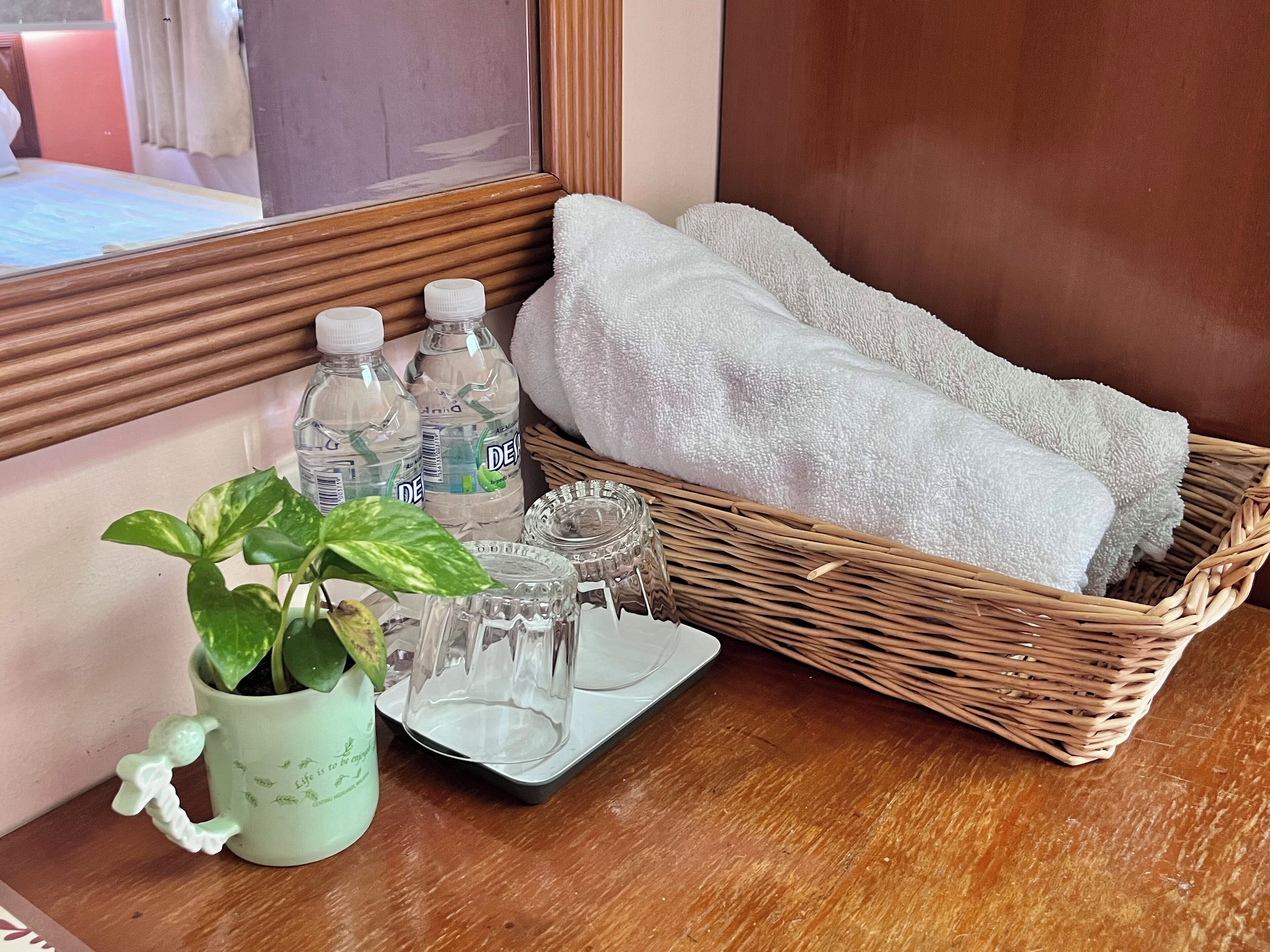 A bathroom vanity with a mirror, a wicker basket containing white towels, two bottles of water, a potted plant, and a tray with glassware.