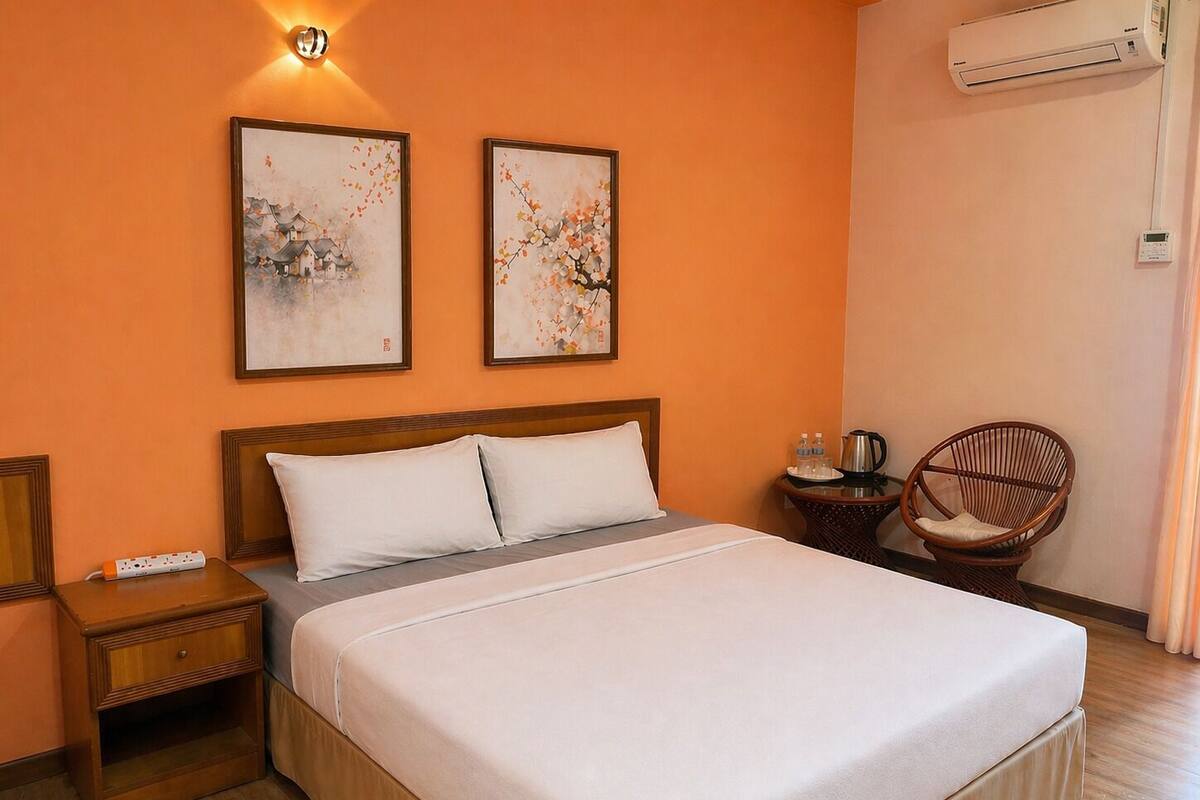 A hotel room with a large bed, two framed artworks on the wall, a bedside table with a lamp, a chair, and an air conditioning unit.