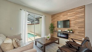 Een moderne woonkamer met een bank, salontafel en lederen fauteuils, een flatscreen televisie bevestigd aan een houten wand, en een schuifdeur die leidt naar een zwembadgedeelte.