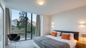 A modern hotel room with a large bed, a chair, and a view of mountains through the window. - 3-BDR Apartment with Panoramic Views & Fireplace (Queenstown)