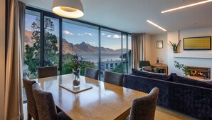 A modern living room with a large window offering a mountain view, a dining area with a wooden table, and a cozy seating area with a fireplace. - 3-BDR Apartment with Panoramic Views & Fireplace (Queenstown)