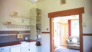 A kitchen with white cabinets, a sink, and shelves with glassware and dishes. There is a wooden door leading to a living area with a sofa and a patterned rug.