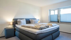 Ein ordentlich bezogenes Bett mit Kissen und Decke, eine Nachttischlampe, ein Fenster mit Jalousien und eine Heizung.