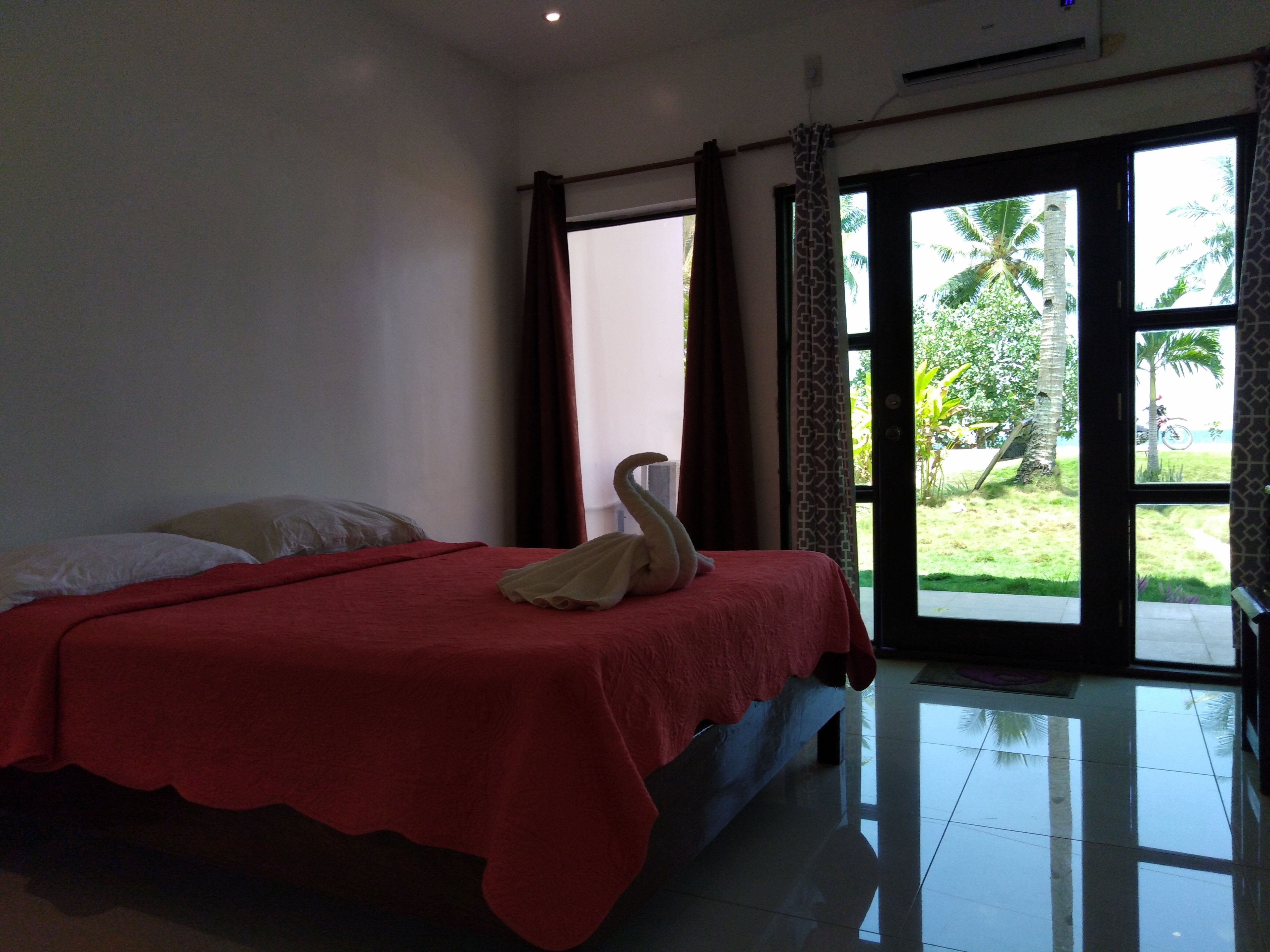 Habitación de hotel con cama, cubrecama rojo, toalla y vista a palmeras por la puerta.