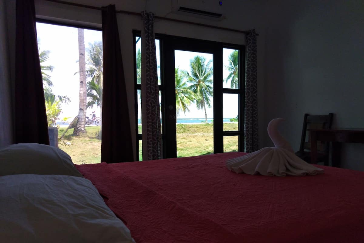 A hotel room with a bed, a view of palm trees and the beach, and a chair by the window.