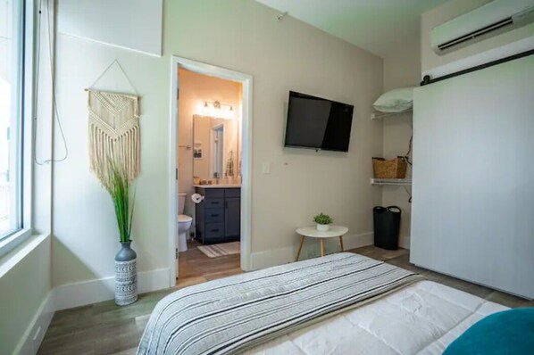 A bedroom with a bed, a TV, a small table with a plant, and a macrame wall hanging.