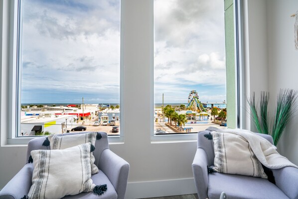 Una habitación con dos sillones grises, una pared blanca y un ventanal con vistas a una playa y un parque de atracciones.