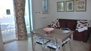 A dining area with a table, chairs, and a bowl. There is a sofa with a patterned cushion and a window with blinds. - Vacation in the House of Dreams (Portoscuso)