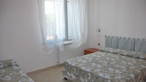 A bedroom with two beds, a window with sheer curtains, a radiator, and a bedside table. - Vacation in the House of Dreams (Portoscuso)