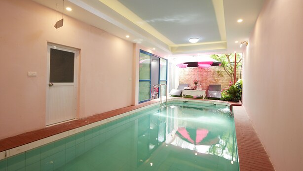 An indoor swimming pool with clear water, a door on the left, and a small seating area with a table and chairs in the background. - Lucky Budda Inn Villa (Hanoi)