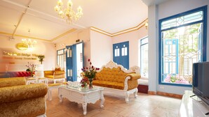 A spacious living room with a large sofa, a coffee table, and a television. There is a chandelier hanging from the ceiling and a window with blue shutters. - Lucky Budda Inn Villa (Hanoi)