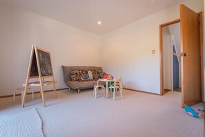 A room with a white carpet, a sofa with a floral cushion, a small table with chairs, and a wooden easel with a chalkboard.