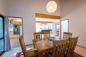 A dining area with a wooden table and chairs, a hanging light fixture, and a view into a kitchen and living space. - Retreat on the Ridge - Lake Ōkareka, Sleeps 8 (Rotorua)