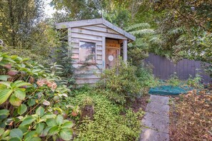 A small wooden structure with a door and window, surrounded by dense foliage and a wooden fence.