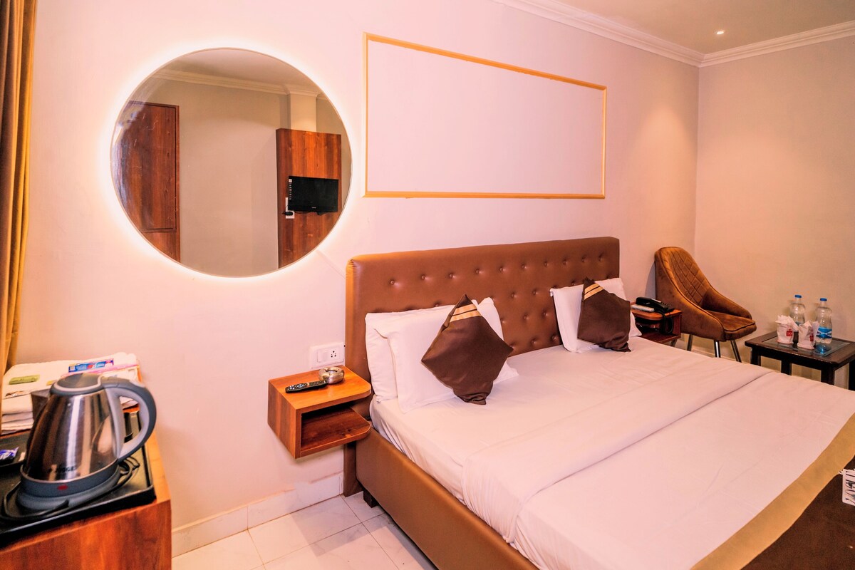 Ein Hotelzimmer mit einem großen Bett, einem runden Spiegel, einem Flachbildfernseher, einem Nachttisch aus Holz, einem Sessel und einem kleinen Tisch mit Wasserkocher und Flaschen.