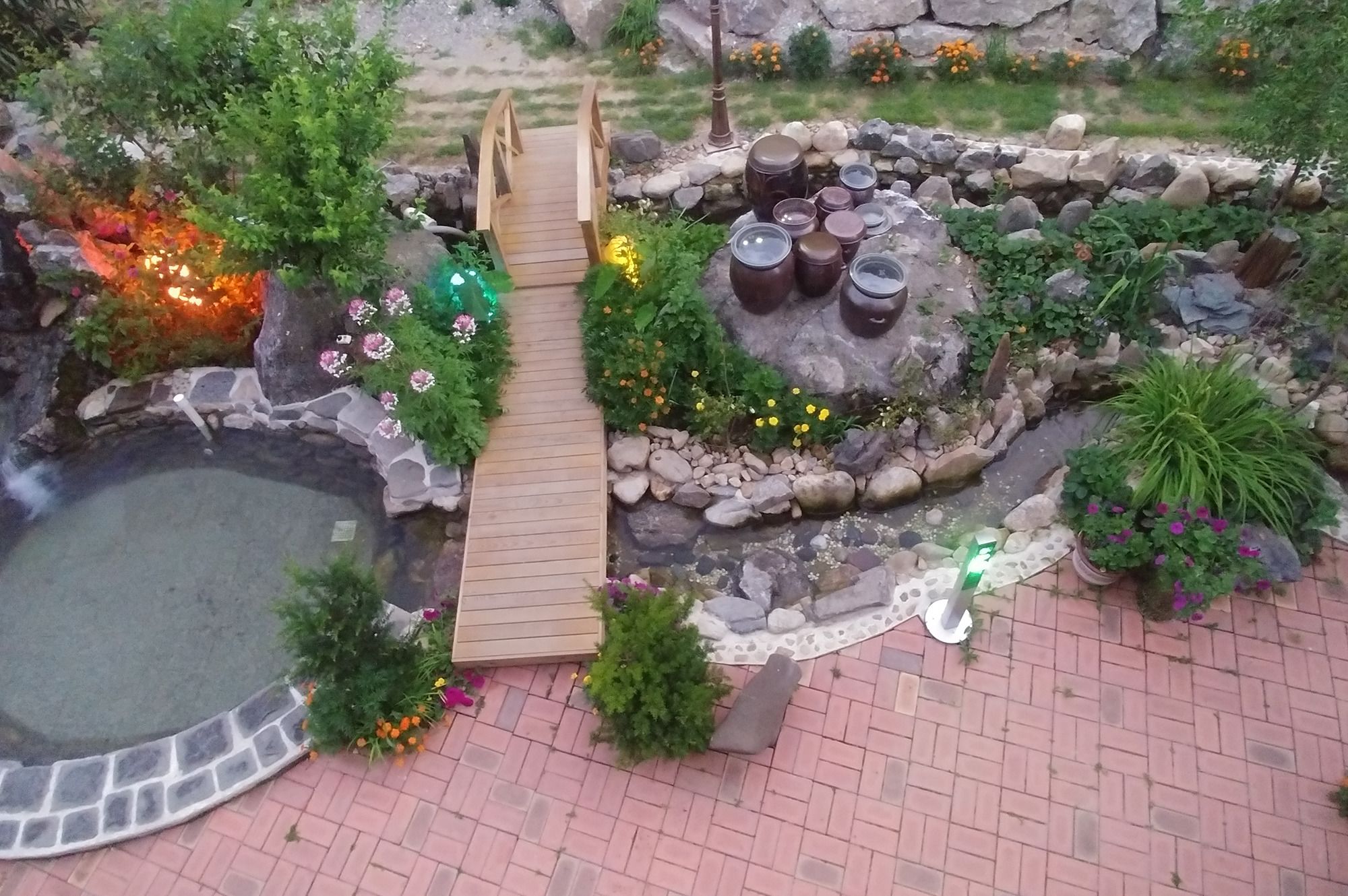 Ein Garten mit einer hölzernen Brücke, einem kleinen Teich und einer Steinmauer mit dekorativen Pflanzgefäßen.