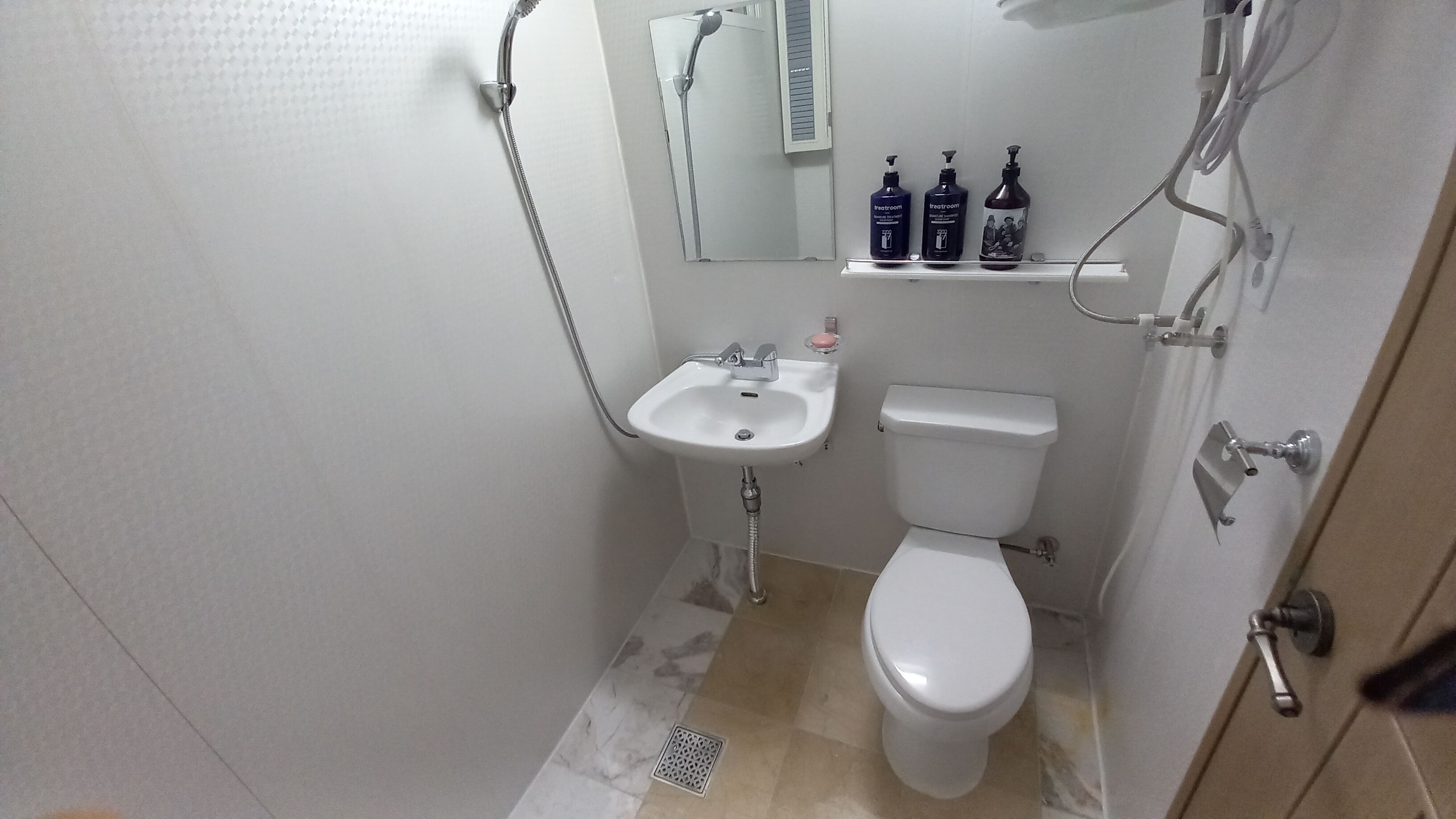 Un baño pequeño con lavamanos, inodoro y ducha blancos. Hay tres botellas de artículos de tocador en un estante sobre el lavamanos.
