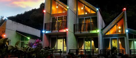 A modern two-story building with balconies and large windows, surrounded by a landscaped garden with red flowers.