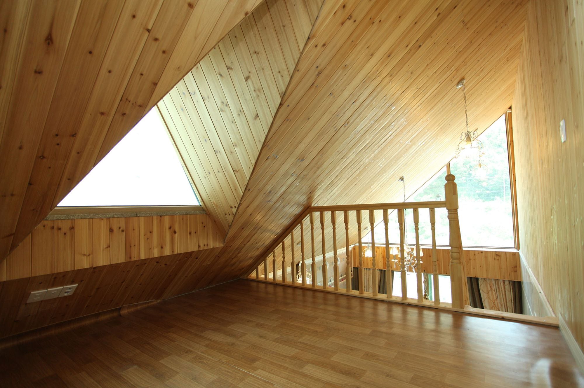 A wooden attic room with a sloped ceiling, wooden railing, and a large window.