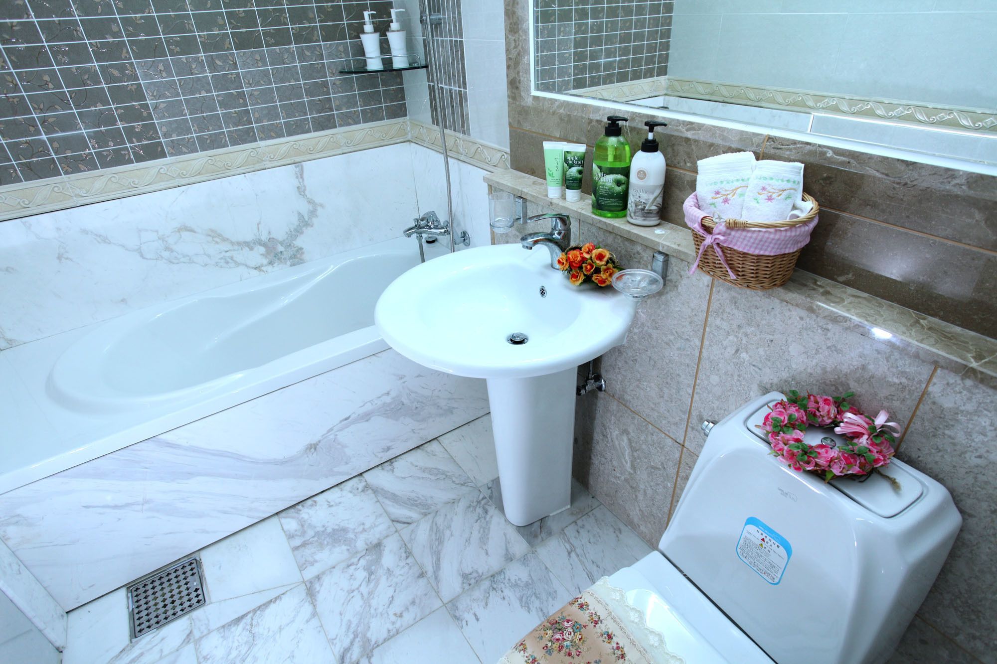 Une salle de bain avec une baignoire, un lavabo et des toilettes. On trouve des articles de toilette sur l’étagère du lavabo et un panier rempli de serviettes accroché au mur.