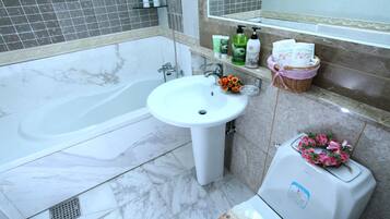 Une salle de bain avec une baignoire, un lavabo et des toilettes. On trouve des articles de toilette sur l’étagère du lavabo et un panier rempli de serviettes accroché au mur.