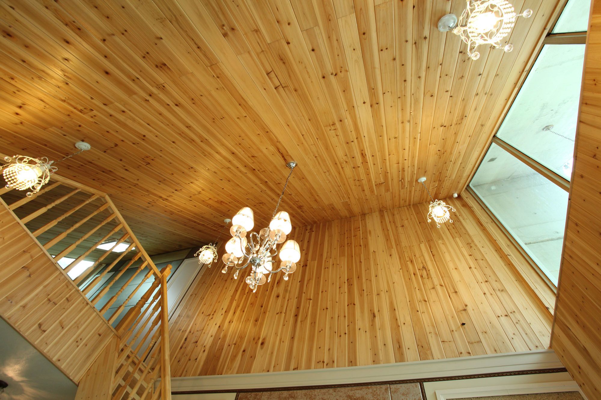 A wooden interior with a wooden staircase, a chandelier, and large windows.