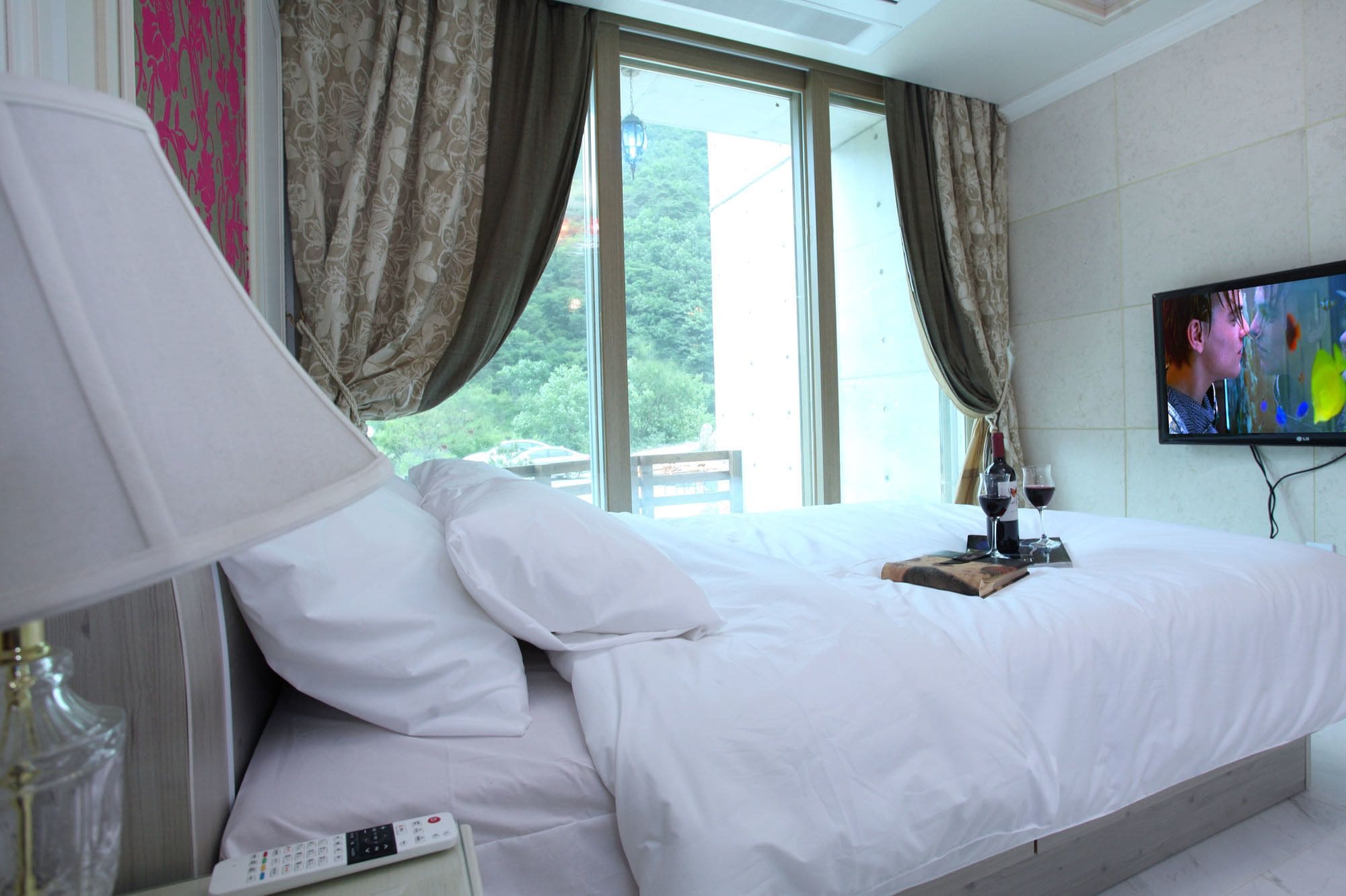 A hotel room with a bed, a TV, a remote control, and a bottle of wine on the nightstand.