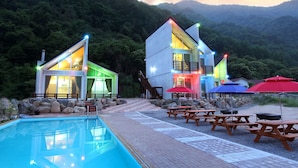Un resort moderno con piscina, mobiliario exterior y vistas a una cadena montañosa boscosa.