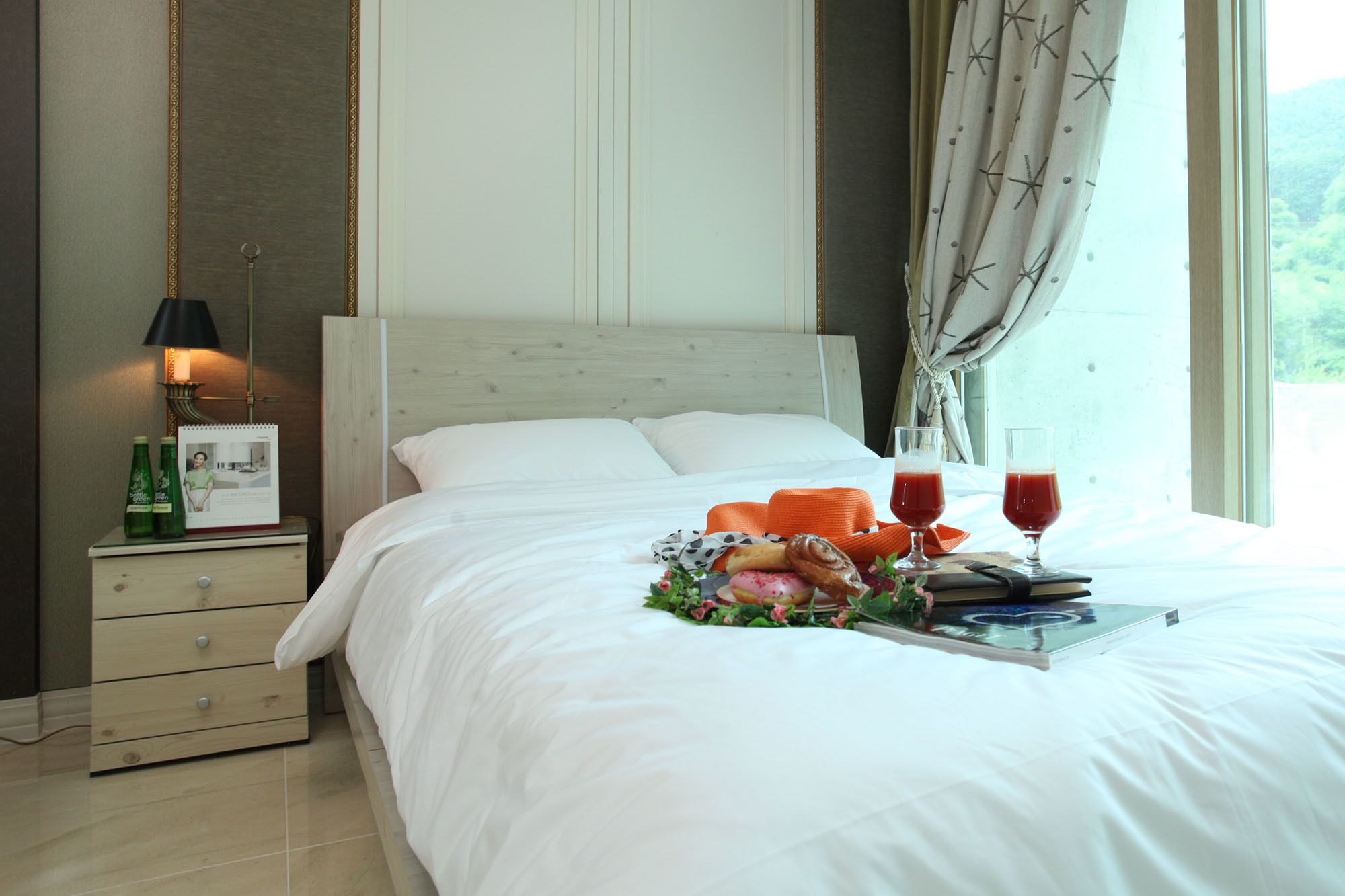A neatly made bed with white linens, a bedside table with a lamp, a tray with pastries, and two glasses of red drink.