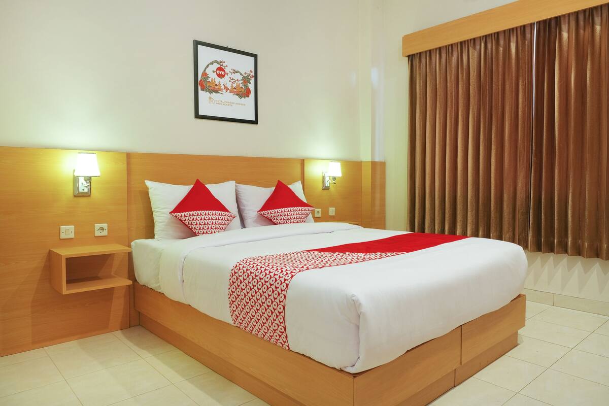 Une chambre d’hôtel avec un lit en bois, du linge de lit blanc, des oreillers rouges, deux tables de chevet avec des lampes et un tableau encadré au mur.