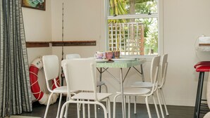 A dining area with a round table and white chairs, a window with a view of greenery, and a painting on the wall.