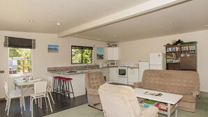 A modern living room with a sofa, coffee table, and dining area. There is a kitchen with white appliances and a bookshelf.