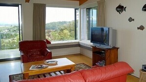 Una sala de estar con un sofá de cuero rojo, una mesa de centro de madera, un televisor sobre un mueble de madera y vistas al exterior a través de grandes ventanales.