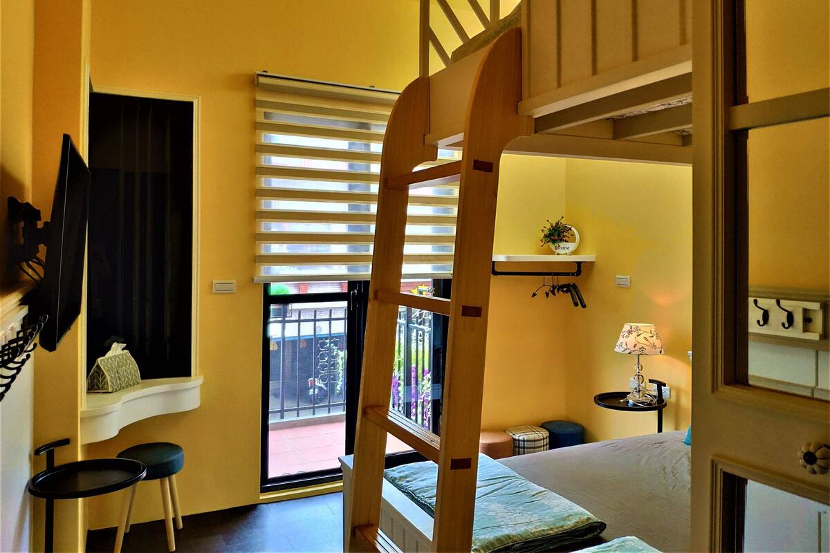 A room with a bunk bed, a window with blinds, a small table with a lamp, and a shelf with a 'home' sign.